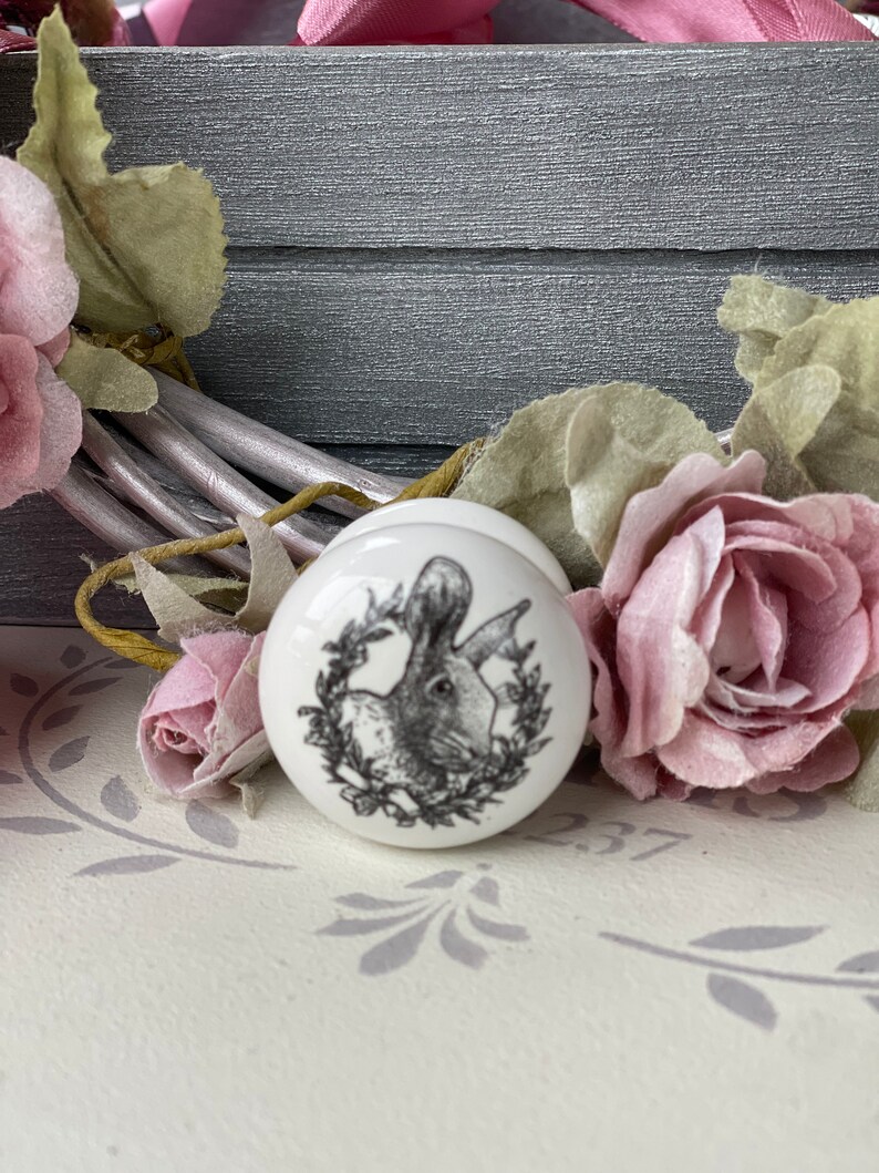 Puede incluir: Pomo de cer&aacute;mica blanco con una imagen en blanco y negro de un conejo en una corona. El pomo est&aacute; sobre una superficie blanca con un patr&oacute;n floral gris.