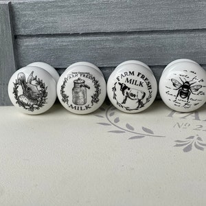 May include: Four white ceramic knobs with black and white illustrations. The knobs feature a rabbit, a milk jug, a cow, and a bee.