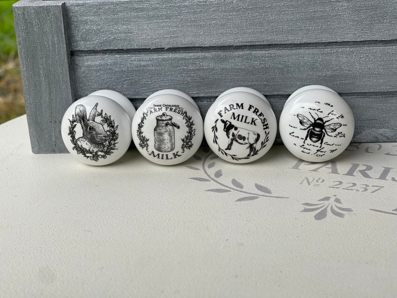 Puede incluir: Cuatro pomos de cer&aacute;mica blanca con ilustraciones en blanco y negro. Los pomos representan un conejo, una jarra de leche, una vaca y una abeja.