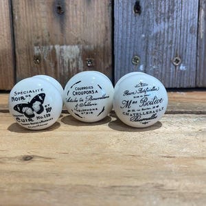 Puede incluir: Tres pomos de cer&aacute;mica blanca con letras y dise&ntilde;os negros. El primer pomo presenta una mariposa y el texto "Specialite Noir de Cuir No. 129". El segundo pomo presenta el texto "Courroies Croupons a Mettre en Boucle a Telephone 9818". El tercer pomo presenta el texto "Fleur d'Anjouchen M.ne Boileve Chatelleraule Damville".