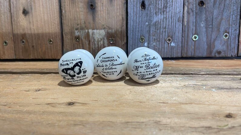 Puede incluir: Tres pomos de cer&aacute;mica blanca con letras y dise&ntilde;os negros. El primer pomo presenta una mariposa y el texto "Specialite Noir de Cuir No. 129". El segundo pomo presenta el texto "Courroies Croupons a Mettre en Boucle a Telephone 9818". El tercer pomo presenta el texto "Fleur d'Anjouchen M.ne Boileve Chatelleraule Damville".