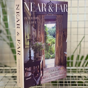 以下が含まれることがあります： 「NEAR & FAR: Interiors I Love」というタイトルのハードカバーの本が表示されています。表紙には、カーテンと屋外の景色がある部屋のインテリアが描かれています。背表紙にもタイトルが表示されています。この本はLisa Fine著、Miguel Flores-Vianna写真です。