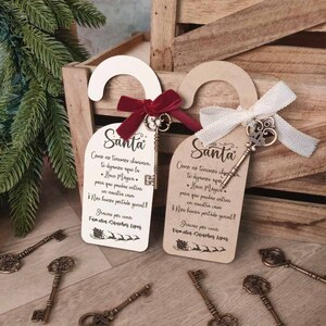 Puede incluir: Dos colgadores de puerta con la palabra "Santa" y texto en español, cada uno con una llave decorativa y una cinta. Varias llaves de estilo antiguo están esparcidas alrededor de los colgadores, con una caja de madera en el fondo.