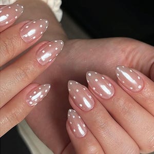Puede incluir: Uñas almendradas, con esmalte nude transparente y lunares blancos. Las uñas tienen un acabado brillante que refleja la luz, creando una estética delicada y femenina. Una manicura de diseño clásico, adecuada para diversas ocasiones.