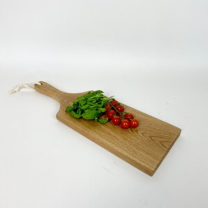 May include: A wooden cutting board with a handle, featuring a light brown color and a natural wood grain. Fresh basil and cherry tomatoes are arranged on the board. A white rope is attached to the handle.