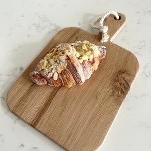 May include: A wooden cutting board with a rope handle, featuring a croissant topped with sliced almonds and powdered sugar.