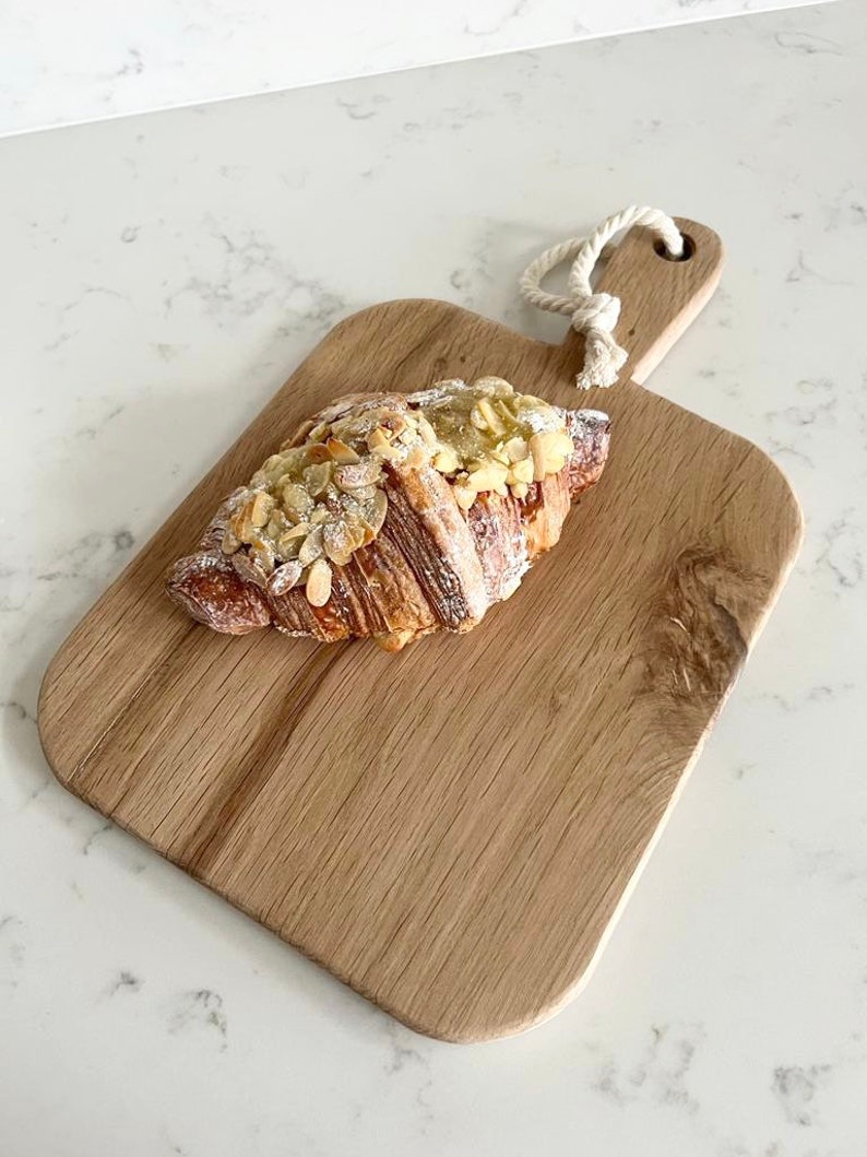 May include: A wooden cutting board with a rope handle, featuring a croissant topped with sliced almonds and powdered sugar.