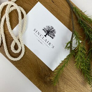 May include: A rectangular wooden cutting board with a light brown finish. A white rope and sprig of green rosemary are on the board. A white card with the text "SINCLAIR'S Hand Crafted Timber - Est.2021-" is also on the board.