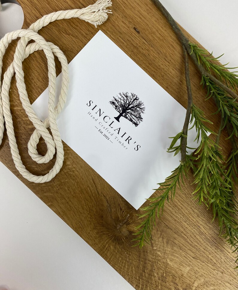May include: A rectangular wooden cutting board with a light brown finish. A white rope and sprig of green rosemary are on the board. A white card with the text "SINCLAIR'S Hand Crafted Timber - Est.2021-" is also on the board.