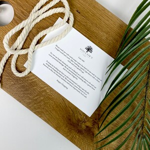 May include: A wooden cutting board with a light-colored rope and a care instruction card. The board has a natural wood grain pattern. A green leaf is in the background. The card has the text "SINCLAIR'S" and care instructions.