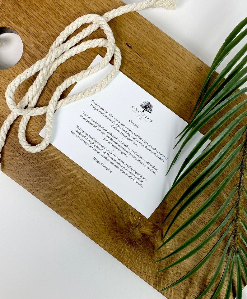 May include: A wooden cutting board with a light-colored rope and a care instruction card. The board has a natural wood grain pattern. A green leaf is in the background. The card has the text "SINCLAIR'S" and care instructions.