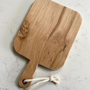 May include: A wooden cutting board with a handle and a rope loop. The board is light brown and has a natural wood grain.
