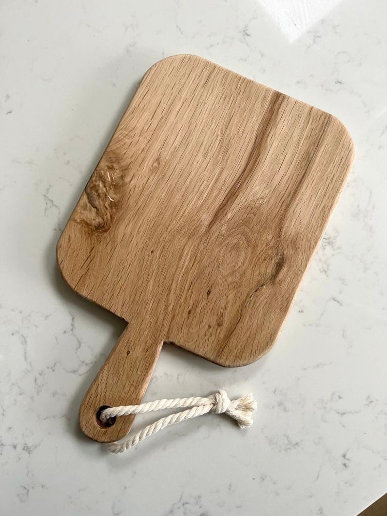 May include: A wooden cutting board with a handle and a rope loop. The board is light brown and has a natural wood grain.