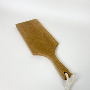 May include: A rectangular wooden serving board with a handle and a hole for hanging. The board is made of light brown wood and has a natural wood grain. A white rope is attached to the handle. The board measures approximately 12 inches long.