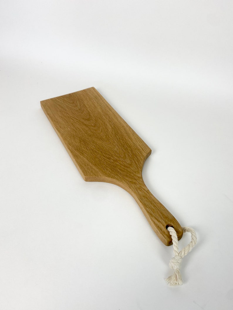 May include: A rectangular wooden serving board with a handle and a hole for hanging. The board is made of light brown wood and has a natural wood grain. A white rope is attached to the handle. The board measures approximately 12 inches long.