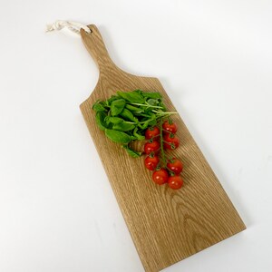 May include: A wooden cutting board with a handle and a white rope loop. Fresh basil and a cluster of red cherry tomatoes rest on the board. The board has a natural wood grain finish.