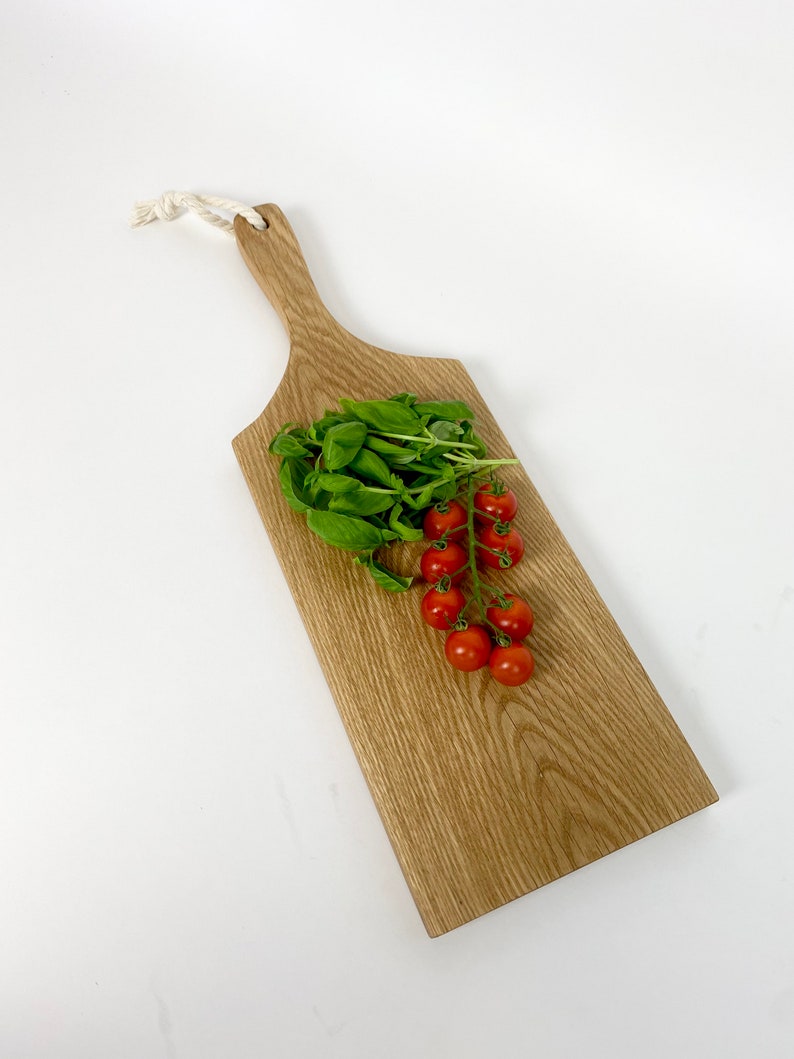 May include: A wooden cutting board with a handle and a white rope loop. Fresh basil and a cluster of red cherry tomatoes rest on the board. The board has a natural wood grain finish.