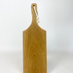 May include: A rectangular wooden cutting board with a handle and a natural wood grain finish. A white rope is attached to the handle for hanging. The board is made of light brown wood.