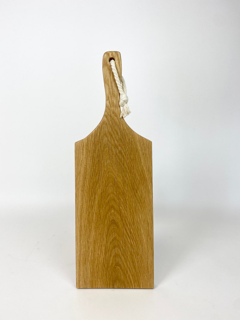May include: A rectangular wooden cutting board with a handle and a natural wood grain finish. A white rope is attached to the handle for hanging. The board is made of light brown wood.