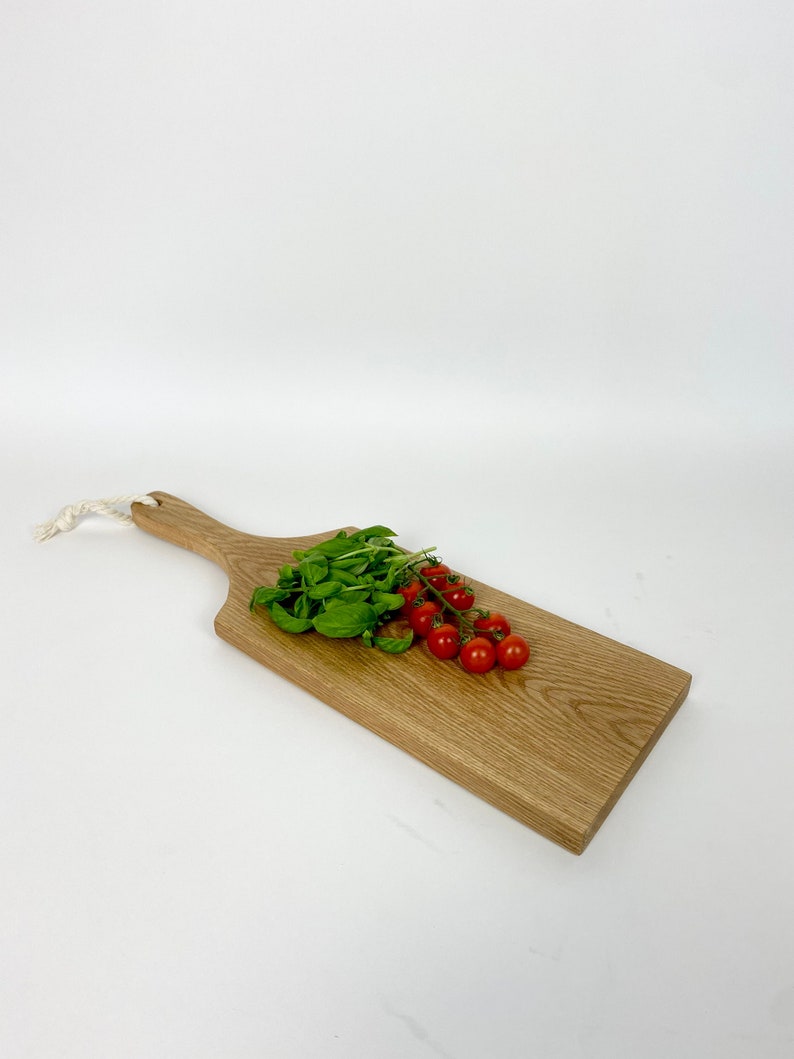 May include: A wooden cutting board with a handle, featuring a light brown color and a natural wood grain. Fresh basil and cherry tomatoes are arranged on the board. A white rope is attached to the handle.