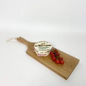 May include: A wooden serving board with a handle, featuring a round cheese wheel labeled "Le Rustique Printemps" and a cluster of red cherry tomatoes. The cheese is wrapped in paper with a red and white checkered pattern.