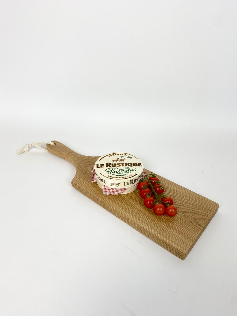 May include: A wooden serving board with a handle, featuring a round cheese wheel labeled "Le Rustique Printemps" and a cluster of red cherry tomatoes. The cheese is wrapped in paper with a red and white checkered pattern.