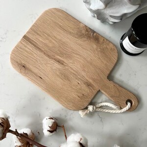 May include: A light brown wooden cutting board with a rounded rectangular shape and a rope handle. The board is on a white marble surface.