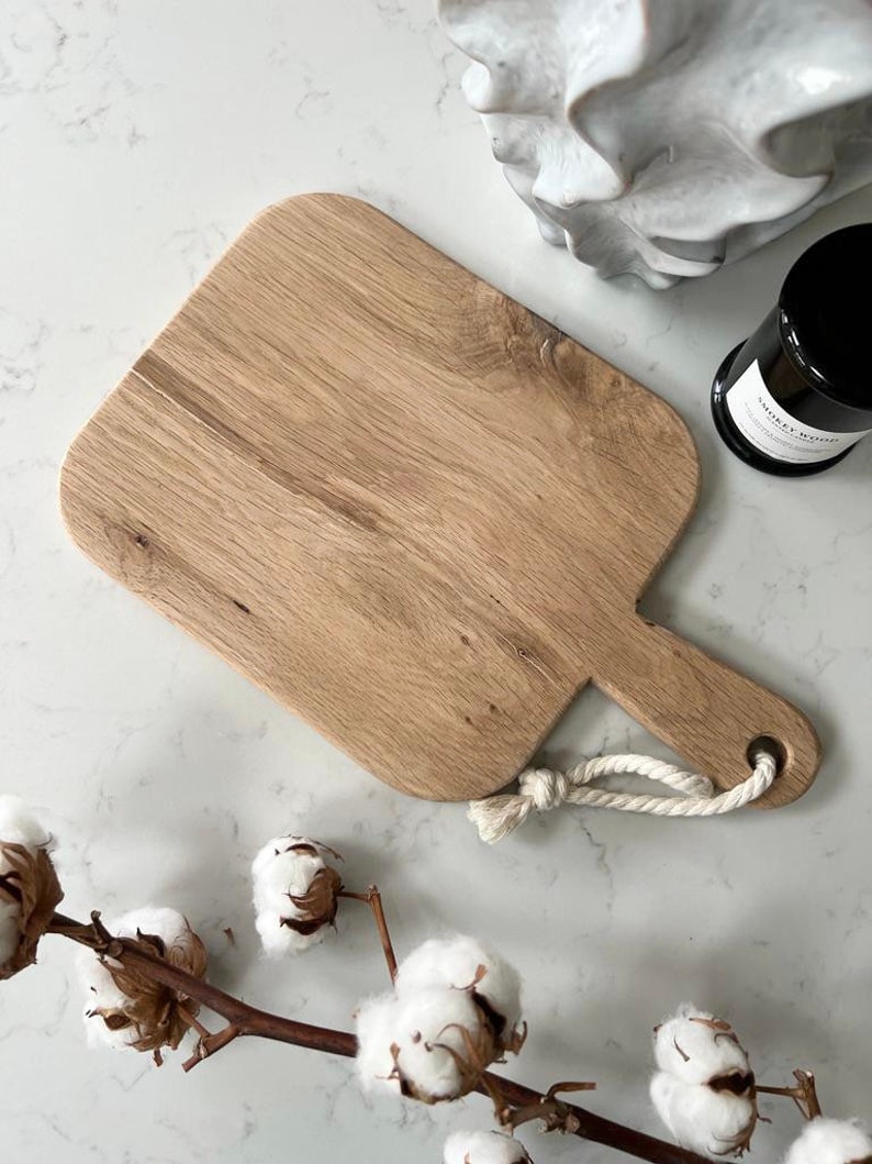 May include: A light brown wooden cutting board with a rounded rectangular shape and a rope handle. The board is on a white marble surface.