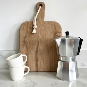 May include: A white countertop with a silver coffee pot, two white ceramic mugs, and a wooden cutting board with a rope handle.