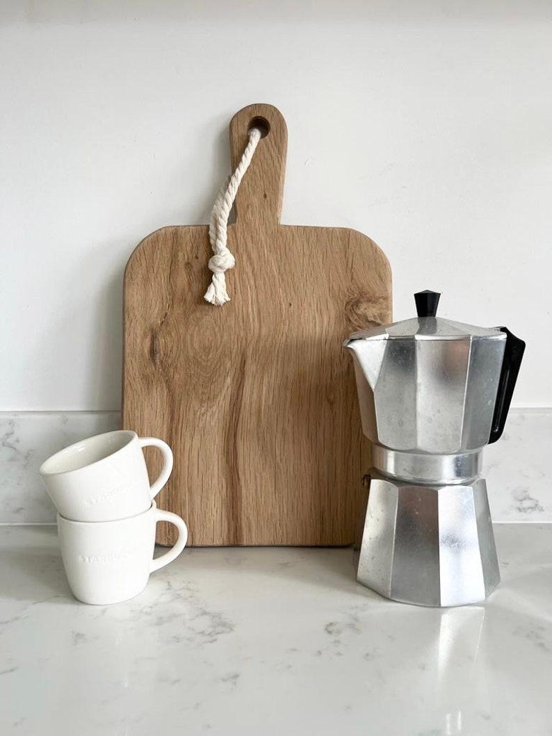 May include: A white countertop with a silver coffee pot, two white ceramic mugs, and a wooden cutting board with a rope handle.
