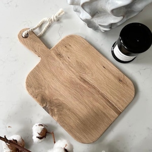 May include: A rectangular wooden cutting board with a rope handle. The board has a natural wood grain and is light brown in color.