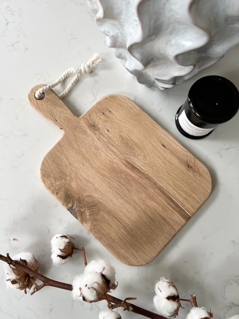 May include: A rectangular wooden cutting board with a rope handle. The board has a natural wood grain and is light brown in color.
