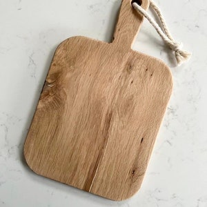 May include: A rectangular wooden cutting board with a natural wood finish and a rope handle. The board has a slightly rounded edge and a smooth surface.