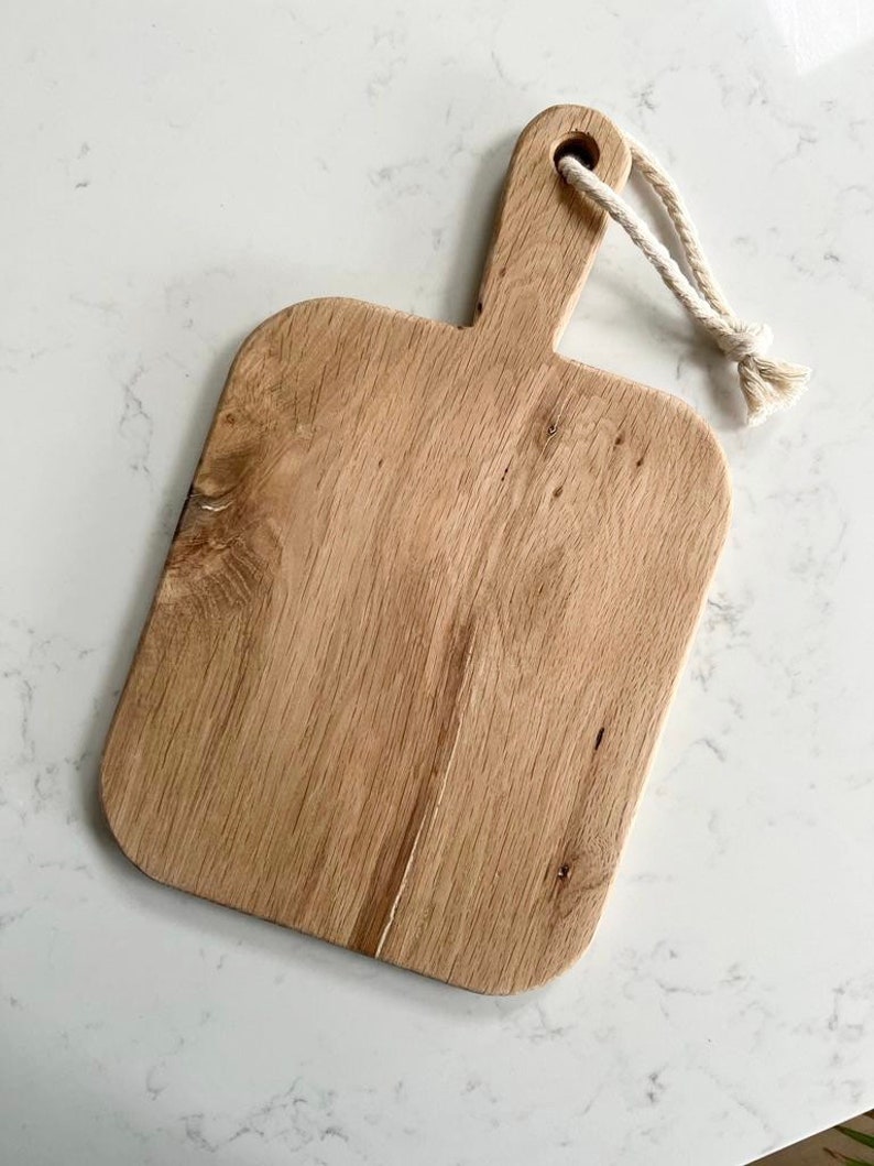 May include: A rectangular wooden cutting board with a natural wood finish and a rope handle. The board has a slightly rounded edge and a smooth surface.