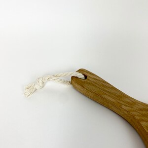 May include: Wooden handled brush with a natural fiber rope loop. The brush has a light brown finish and a curved handle. The rope is white and knotted. The background is white.