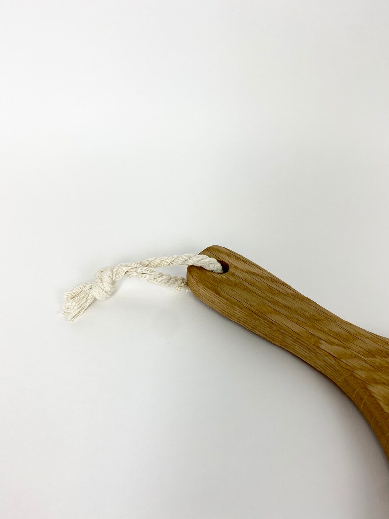 May include: Wooden handled brush with a natural fiber rope loop. The brush has a light brown finish and a curved handle. The rope is white and knotted. The background is white.