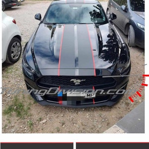 May include: Black sports car featuring grey racing stripes with red accent lines. The car has a front number plate and is parked outside. The image shows the front of the car, with other vehicles and trees in the background.