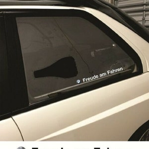 Könnte beinhalten: Ein weißer BMW mit einer zerbrochenen Heckscheibe. Die zerbrochene Scheibe hat ein schwarzes Loch in Form einer Flasche. Das Auto hat einen Aufkleber auf der Scheibe, der "Freude am Fahren" mit dem BMW-Logo sagt.