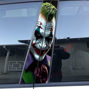 May include: A car window decal featuring a close-up of the Joker's face. The Joker is wearing green hair, white makeup, and a purple suit. The decal is placed on the car's window pillar.