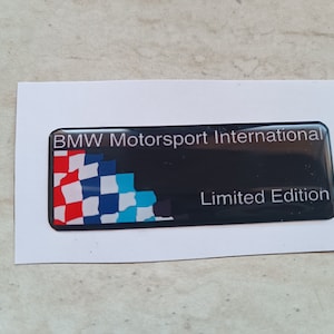 May include: A black rectangular emblem with the text "BMW Motorsport International Limited Edition." The emblem features a checkered flag design in red, white, and blue. The emblem is on a white background.