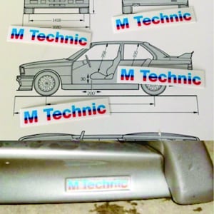 May include: A close-up of a silver car part with a blue, red, and white "M Technic" logo. The logo is on a silver surface with a slight texture.