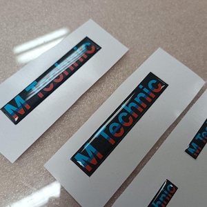 Könnte beinhalten: Drei weiße rechteckige Aufkleber mit schwarzem Hintergrund und dem Text "M Technic" in blauen und roten Buchstaben. Die Aufkleber befinden sich auf einer weißen Oberfläche.