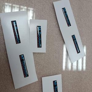 May include: Several white rectangular stickers with the text "M Technic" in a vertical format. The text is in a dark blue font with a gradient of red and blue. The stickers are scattered on a light brown surface.