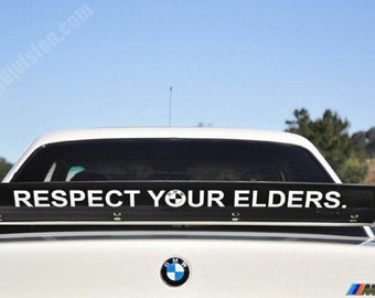 Puede incluir: Un BMW M3 blanco con un alerón trasero negro que dice "RESPECT YOUR ELDERS." El coche tiene un logotipo BMW azul y blanco en la parte trasera.