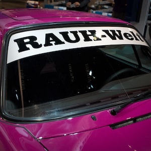 Puede incluir: Un coche rosa con una pancarta negra en el parabrisas que dice "RAUH-Welt".