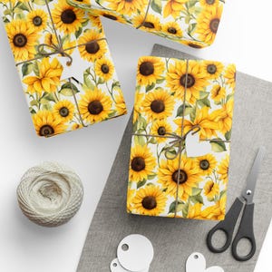 May include: Gift-wrapped packages with a vibrant sunflower pattern. Each package is tied with twine and a heart-shaped tag. A spool of twine, scissors, and blank tags are also visible, suggesting a crafting or gift-giving theme.