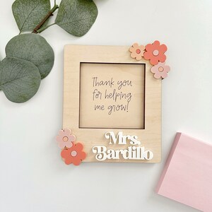 May include: A wooden picture frame with a white background and the text "Thank you for helping me grow!" inside. The frame is decorated with pink and orange flowers and the name "Mrs. Bardillo" is written in white on the bottom.
