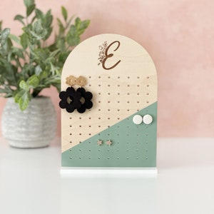 May include: Wooden earring holder with a light green and natural wood finish. The holder has a rounded top with the letter "E" and floral design engraved on it. The holder has a variety of holes for displaying earrings. There are black flower earrings, white round earrings, and gold star earrings displayed on the holder.