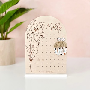 May include: Wooden earring holder with a daffodil design and the name "Molly" engraved on it. The holder has multiple holes for storing earrings. The bottom of the holder reads "March | Daffodil".