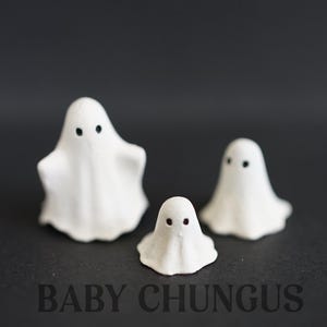 May include: Three small, white ghost figurines with black dot eyes are arranged on a dark surface. The ghosts vary in size, with the largest in the back and the smallest in the front. The text "BABY CHUNGUS" is printed in dark letters.